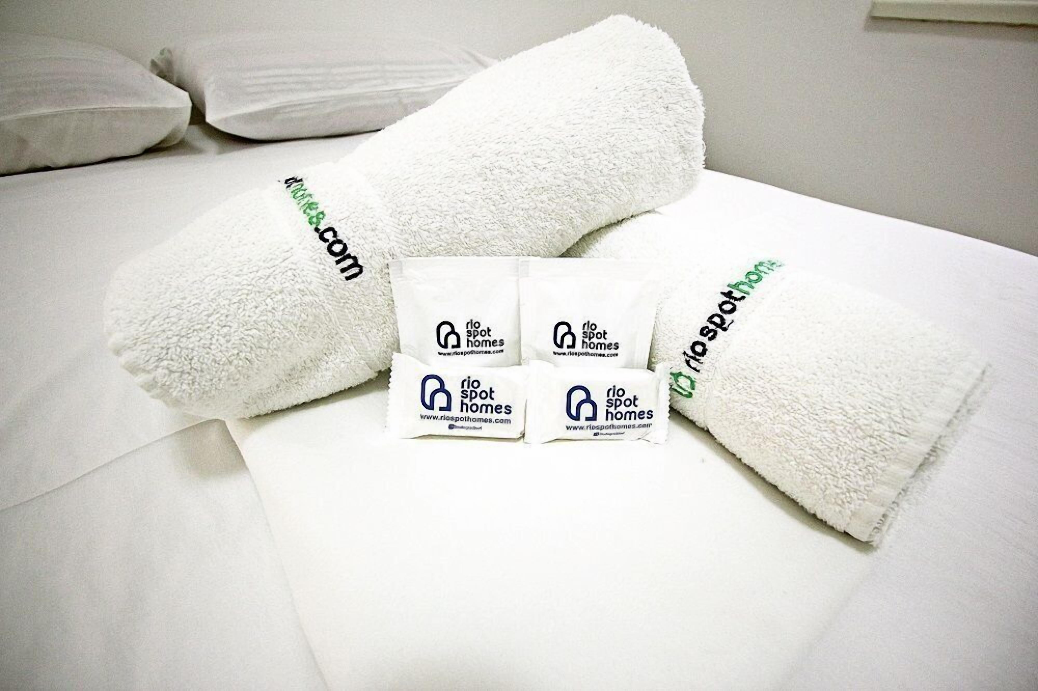 A neatly made bed with white linens and two rolled towels with the logo and website of Rio Spot Homes.