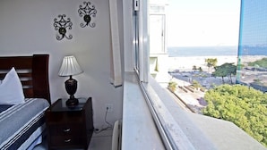 Une chambre à coucher comprenant un lit, une table de chevet, une lampe et des éléments décoratifs sur les murs. Une fenêtre offre une vue sur le paysage urbain et l’océan.