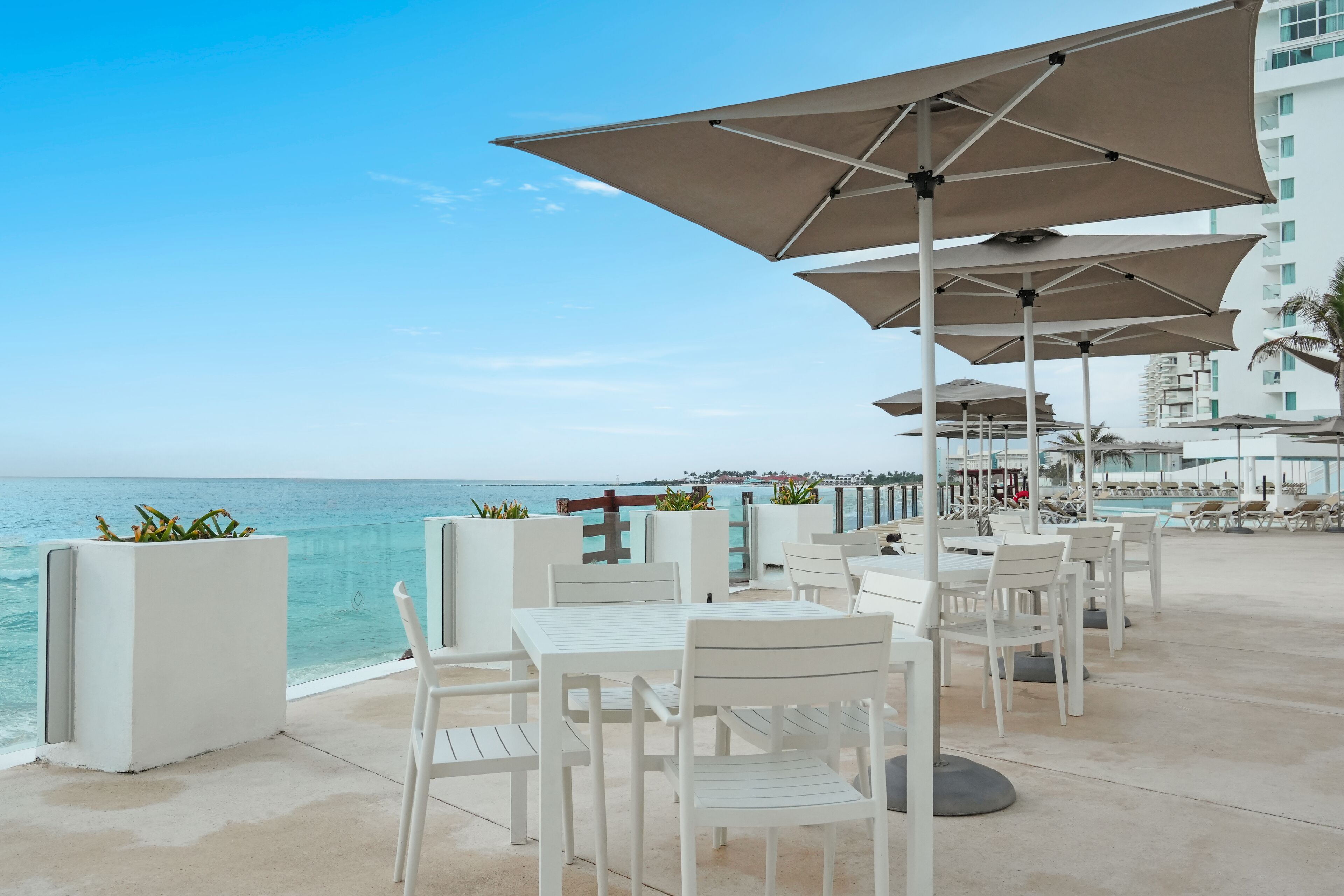 A seaside patio with white tables and chairs, beige umbrellas, and a view of the ocean.
