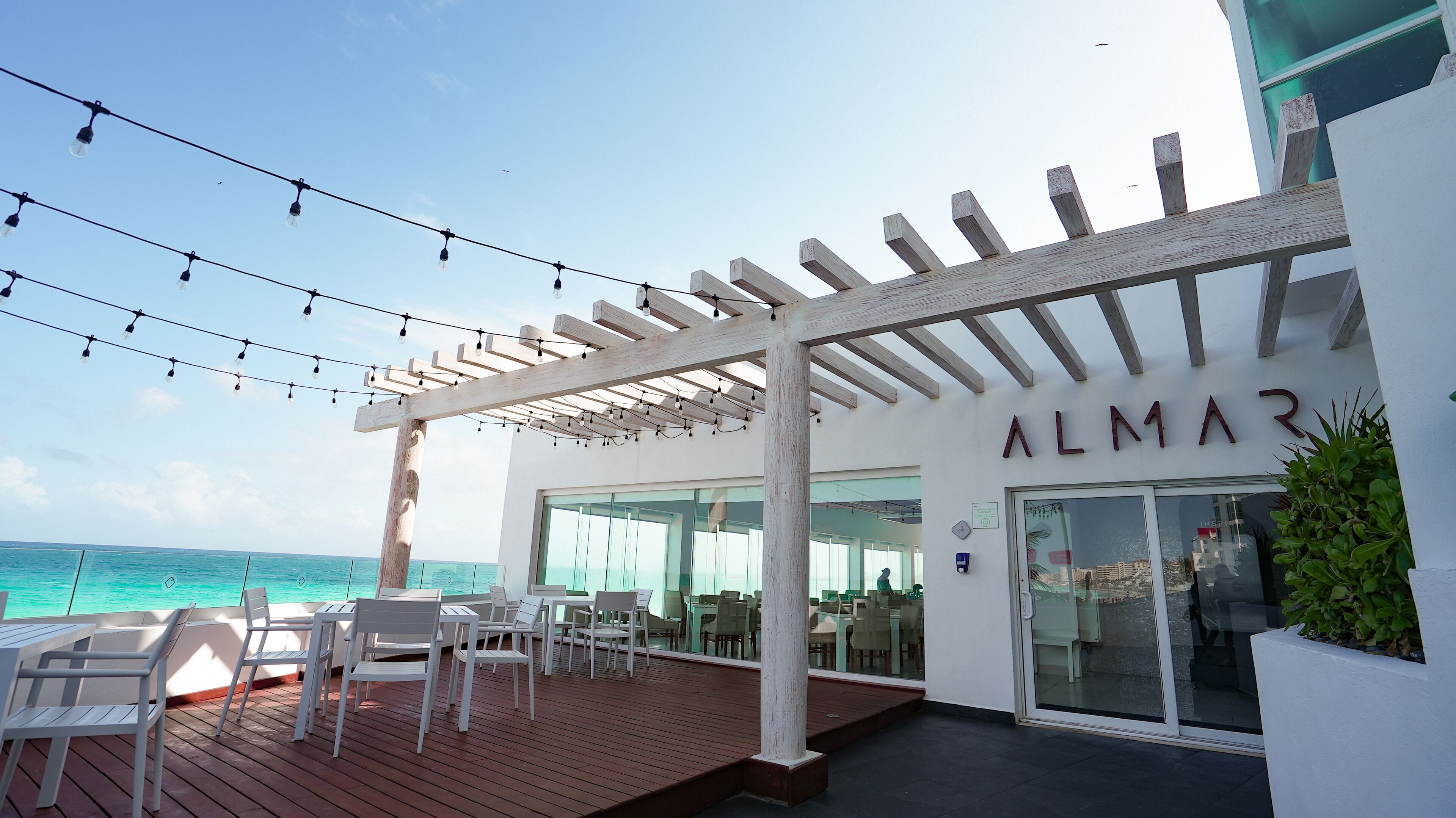 A restaurant with outdoor seating overlooking the ocean, a pergola with string lights, and a sign reading "ALMAR".