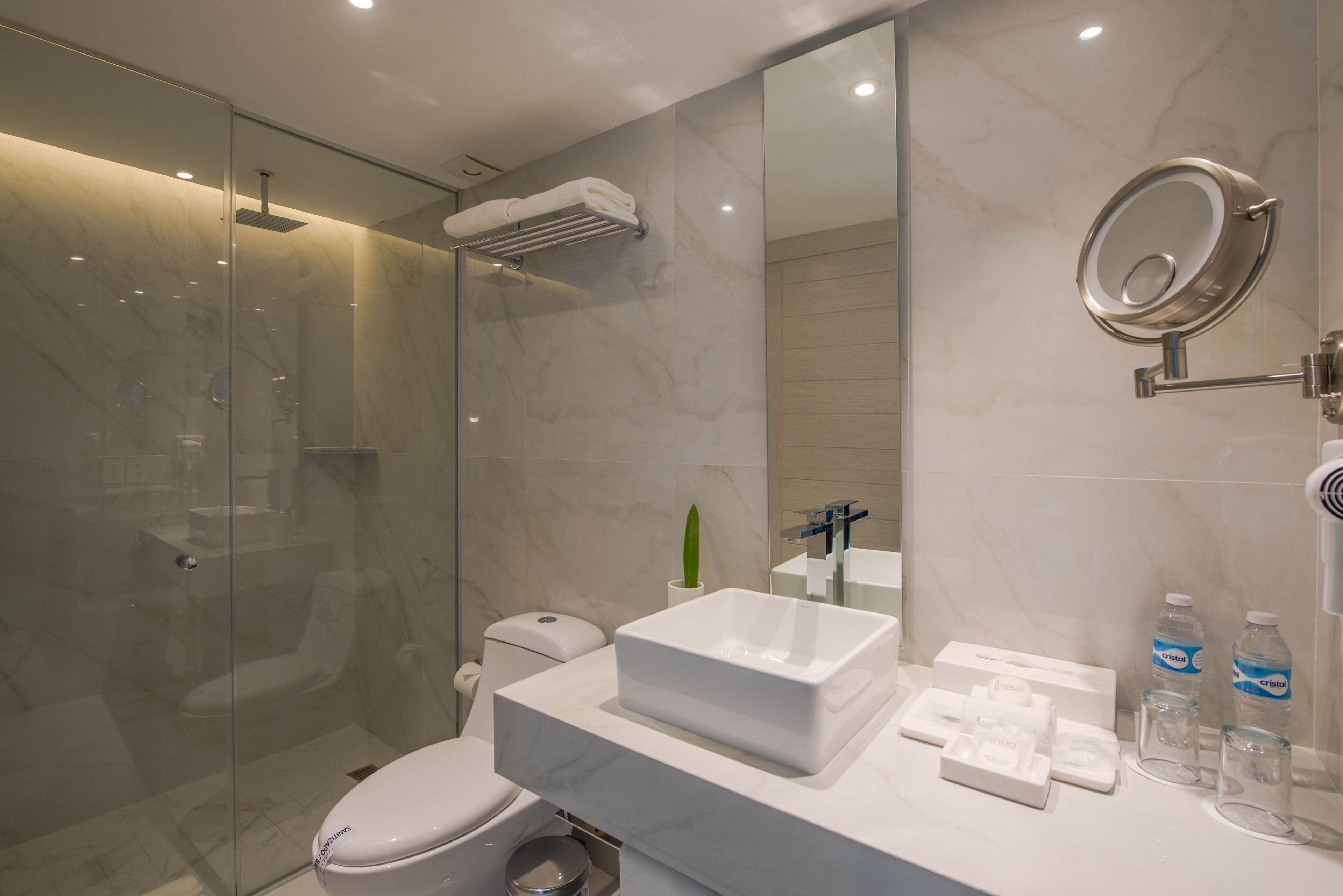 Un baño moderno con ducha envuelta en vidrio, inodoro blanco, espejo grande y encimera con artículos de aseo.