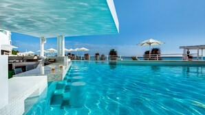 Una piscina azul cristalina con vista al océano, sombrillas blancas y sillas de descanso.