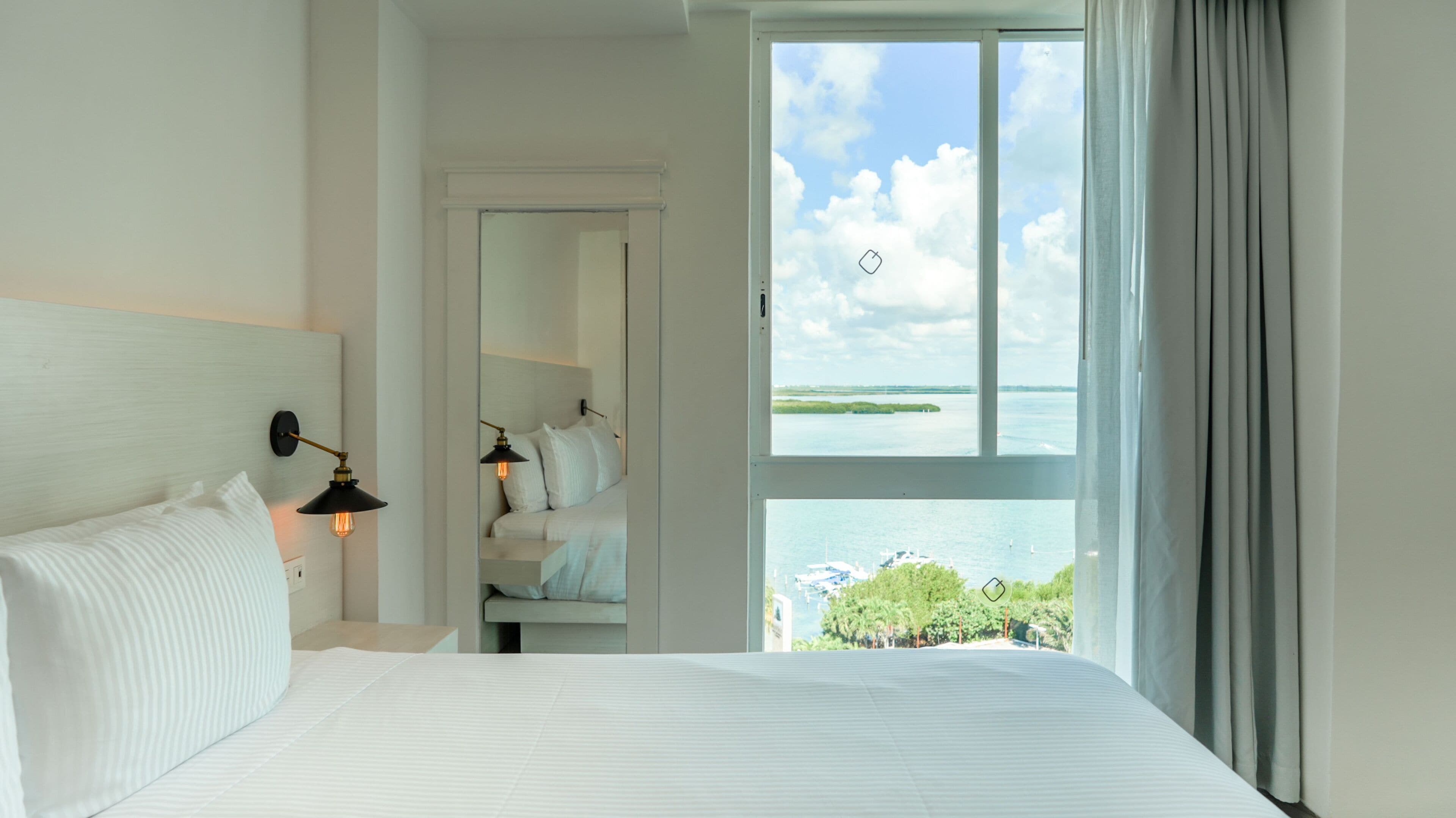 A hotel room with a bed, a nightstand, a mirror, and a view of the sea through a large window.