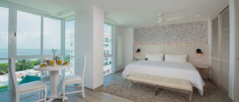 A bedroom with a large bed, a bedside table, a chair, a small table with a plant, and a view of the ocean.