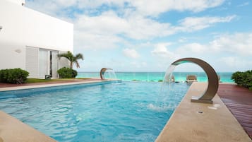 Una piscina con fuente, vista al océano y un edificio blanco con contraventanas.