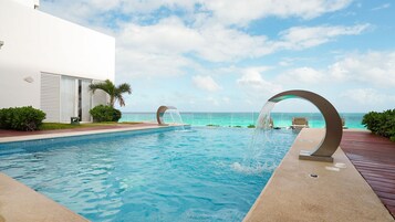 Una piscina con fuente, vista al océano y un edificio blanco con contraventanas.