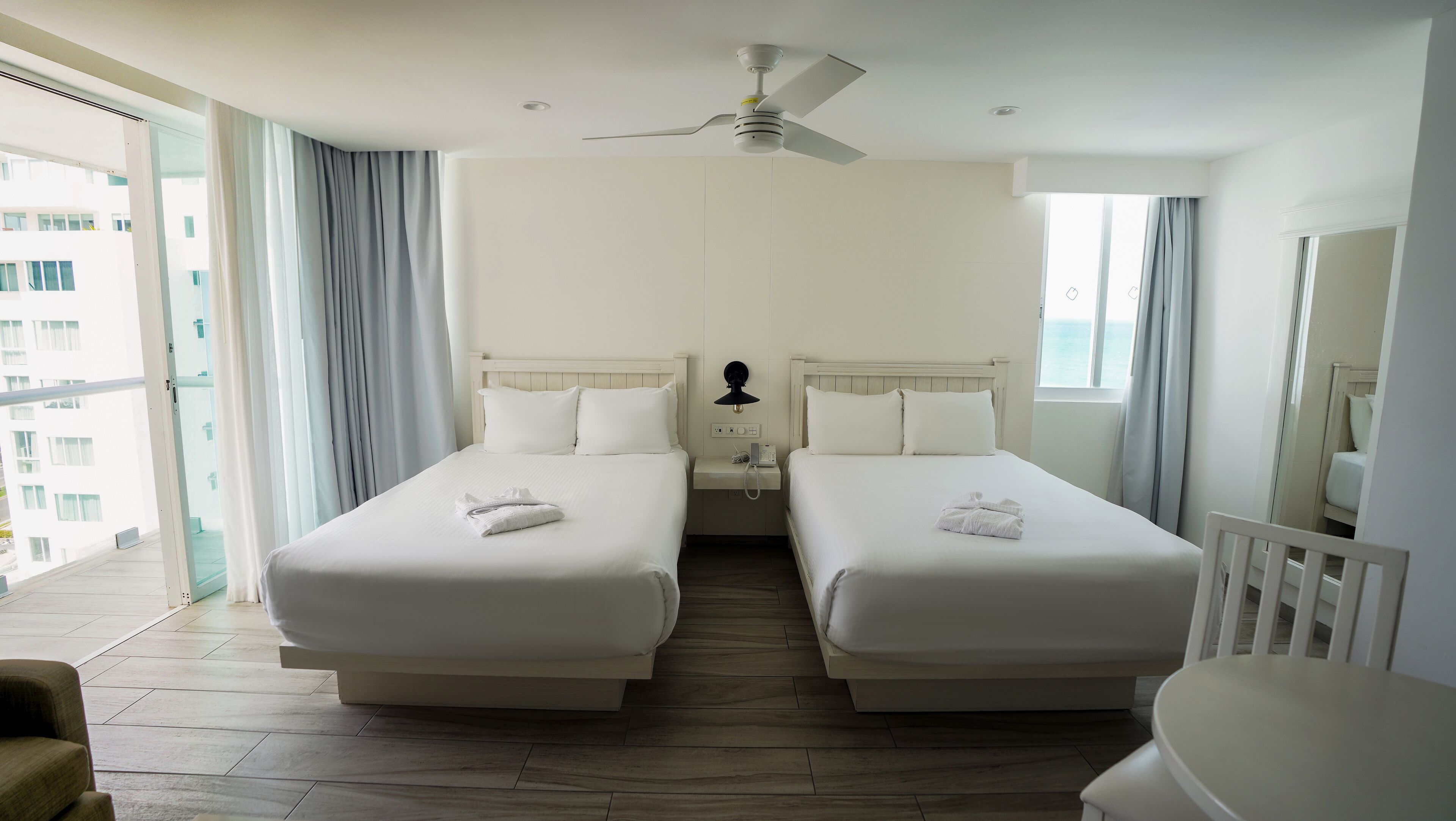 A hotel room with two beds, a ceiling fan, a bedside table with a lamp, and a view of the sea.
