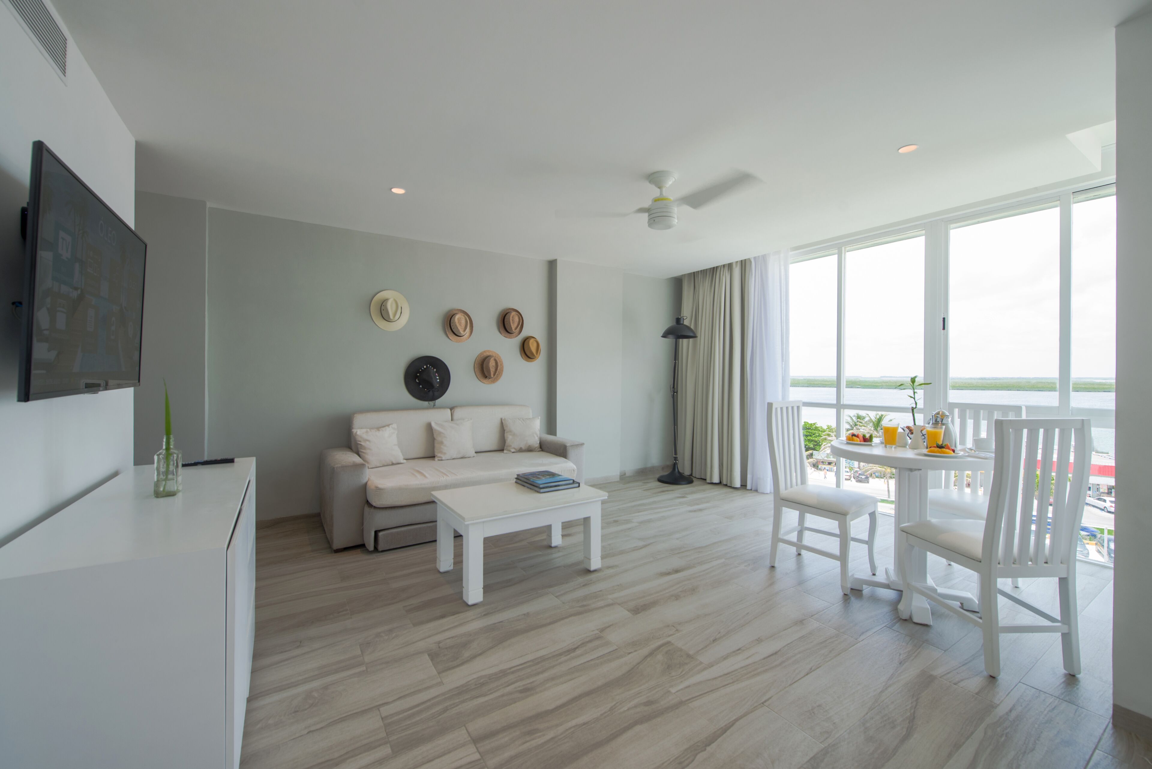 A modern living room with a white sofa, a coffee table, and a dining area with white chairs. There is a ceiling fan, a flat-screen TV mounted on the wall, and a view of the sea through large windows.