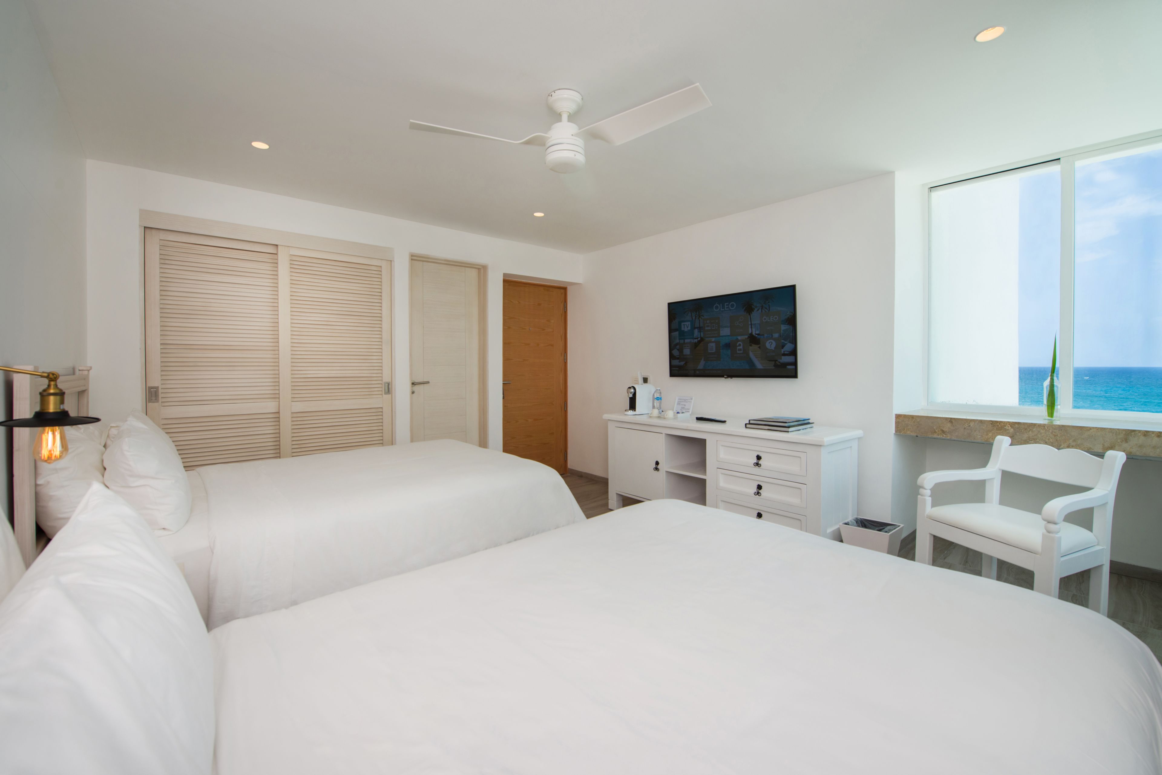 Une chambre d ’hôtel avec deux lits, un bureau, une chaise, un ventilateur de plafond et une grande fenêtre donnant sur l’océan.