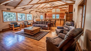 A spacious living area with wooden beams, large windows, a sofa, a leather chair, and a wooden chest of drawers. - Colorado Bear Creek Cabins (Evergreen)