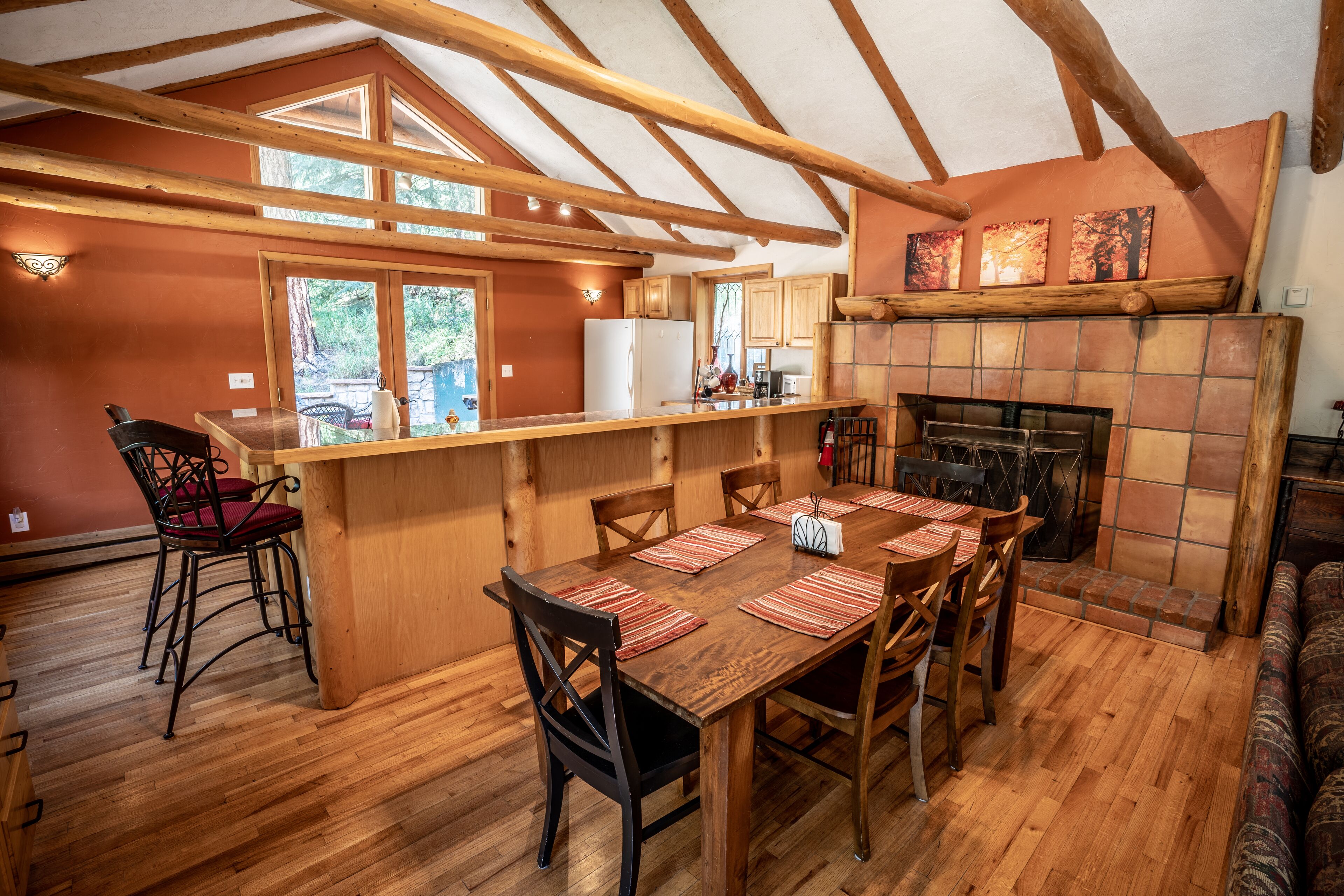 A rustic kitchen with wooden beams, a dining area with a long table and chairs, and a fireplace.