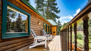 A log cabin with a wooden deck, two white chairs, and a view of the surrounding forest. - Colorado Bear Creek Cabins (Evergreen)