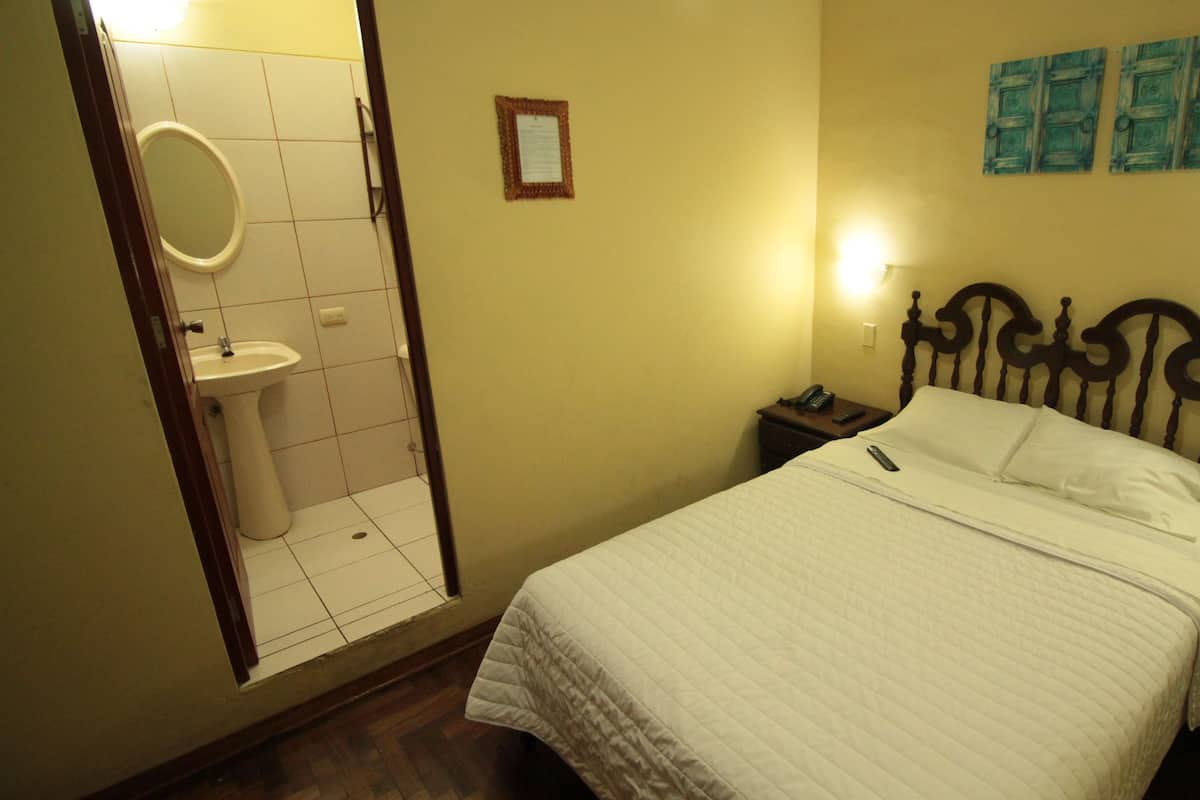 A hotel room with a bed, a bedside table with a phone, a nightstand, a wall-mounted light, a mirror, and a toilet.
