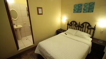 A hotel room with a bed, a bedside table with a phone, a nightstand, a wall-mounted light, a mirror, and a toilet.