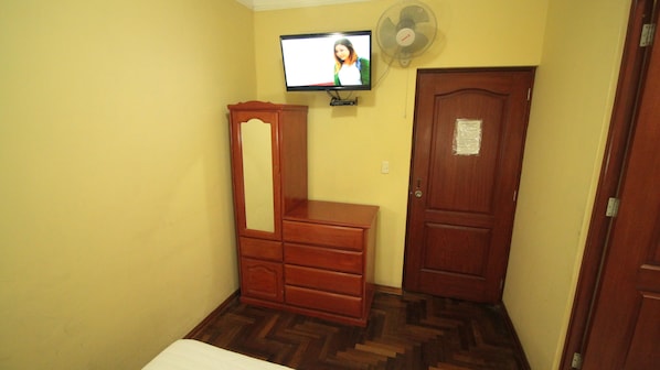 Una habitación pequeña con un aparador de madera, un televisor montado en la pared, una puerta de madera y un ventilador de pie.