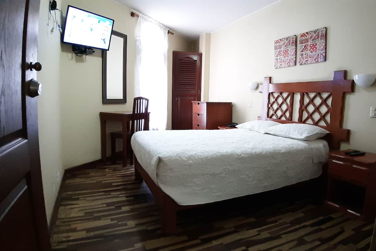 A bedroom with a bed, a desk, a chair, a wardrobe, and a television on the wall.