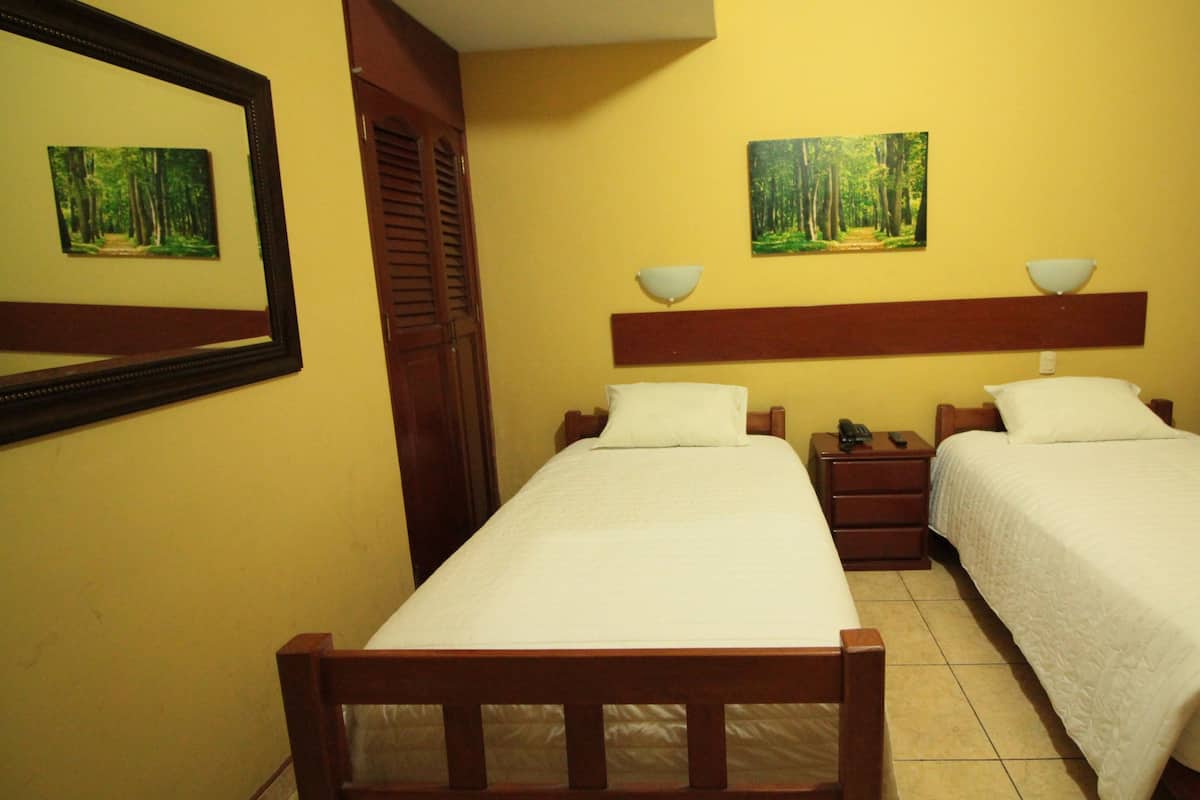 A hotel room with two single beds, wooden headboards, a nightstand with a lamp, and a painting of a forest on the wall.