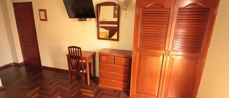 A room with a wooden wardrobe, a desk with a chair, a television, and a wall-mounted fan.