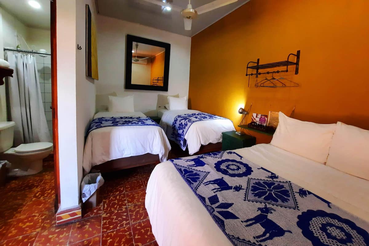 Habitación de hotel con dos camas, una cama grande, ventilador de techo, espejo, mesita de noche con lámpara y cortina de ducha.