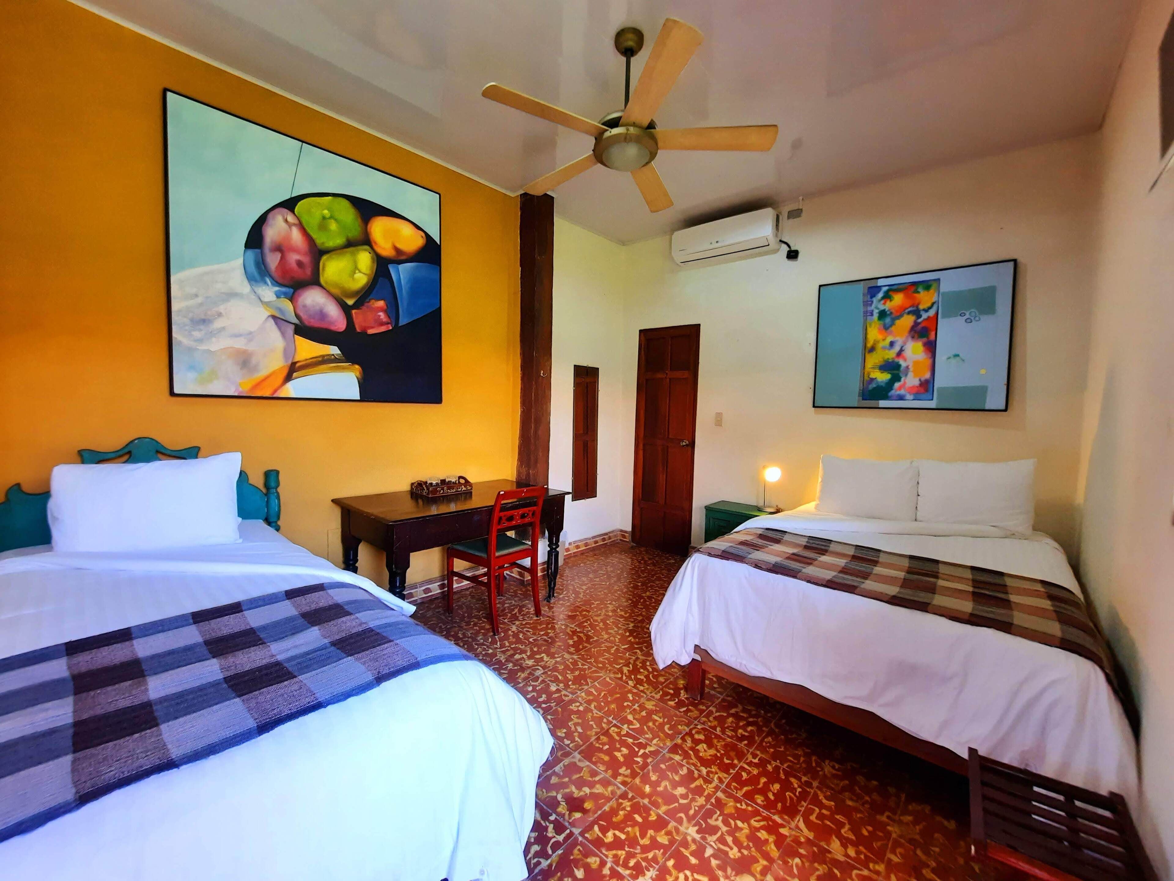 A hotel room with two beds, a desk, a chair, a ceiling fan, and artwork on the walls.