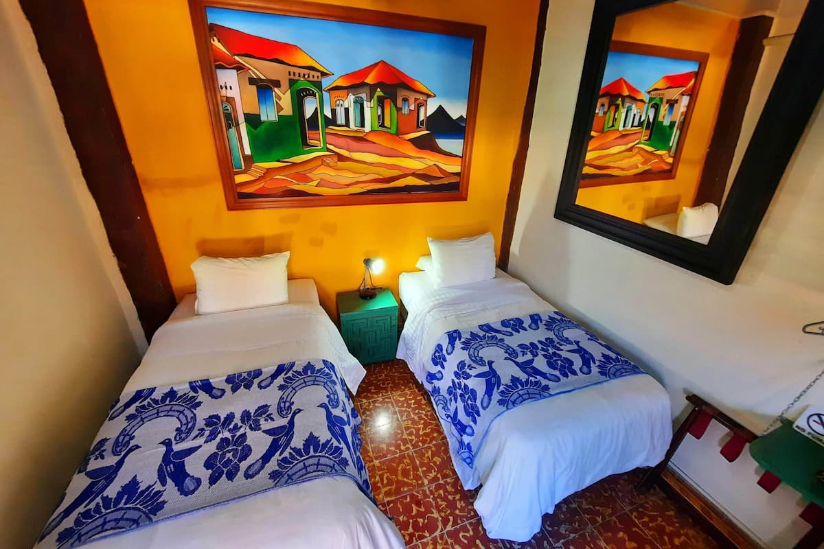 Habitación de hotel con dos camas, cada una con ropa de cama blanca y motivos florales azules. Encima de las camas hay un cuadro que representa una escena de pueblo, y un espejo en la pared.