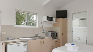A compact hotel room with a kitchenette, a bed with white linens, a television mounted above the microwave, and a window providing natural light.