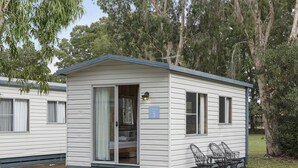A small, single-story mobile home with a blue roof and white siding. It has a sliding glass door leading to a bedroom with a bed and a nightstand. There is a small porch with a table and chairs, and a sign indicating the unit number 3.