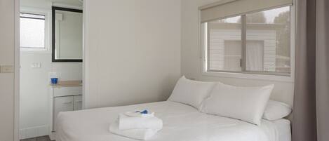 A neatly made bed with white linens and pillows, a small bedside table with a blue cup, and a window with a white blind.