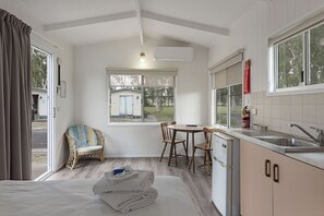 Habitación pequeña con cama, mesa de comedor con sillas, kitchenette y ventana con vistas a un espacio exterior.