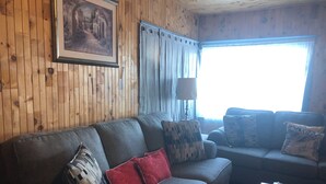 A cozy living room with a wooden paneled wall, a framed picture, a sofa with patterned cushions, and a coffee table with remote controls. - Beautiful Seaside Cottage (Digby)