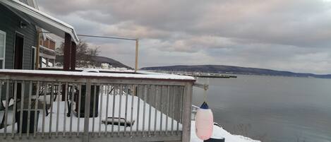 A snow-covered deck with a railing, a dock extending into a calm body of water, and a distant shoreline with hills.