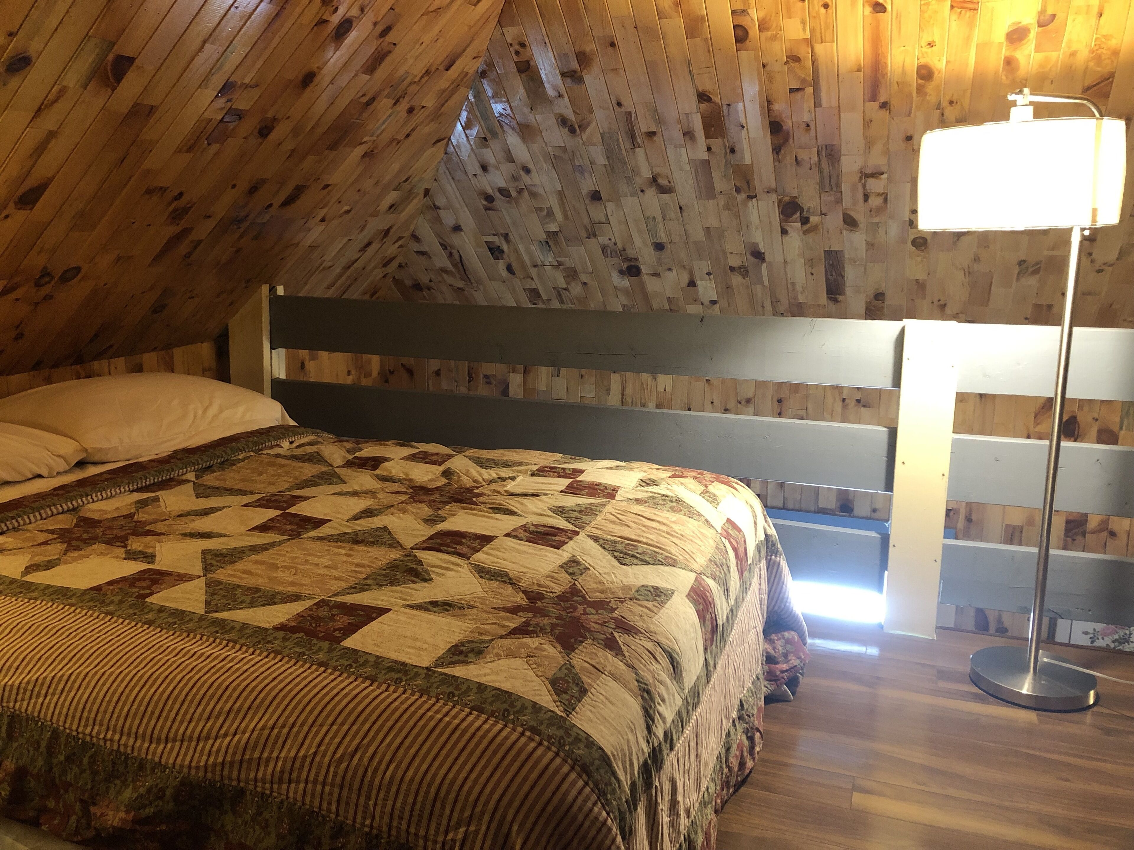 Un dormitorio con techo inclinado de madera, una cama con colcha, una mesita de noche y una lámpara de pie.