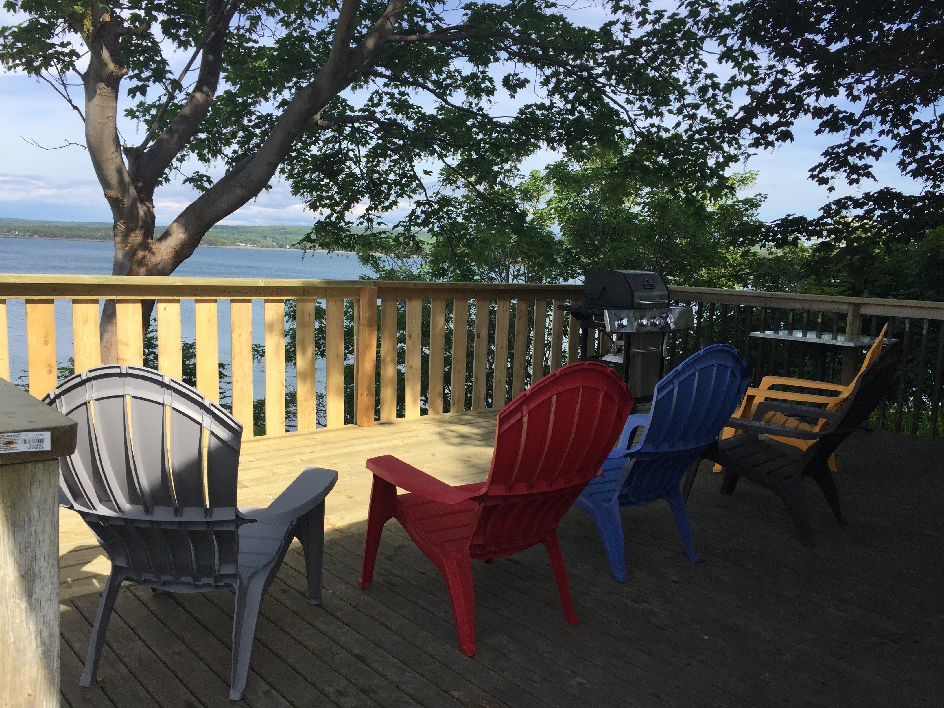 A wooden deck with a view of a lake, featuring a grill and three Adirondack chairs.