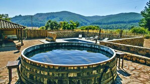 Une piscine extérieure rustique, avec des murs en pierre et des bancs en bois, entourée d’une terrasse en pierre et offrant une vue sur une chaîne de montagnes boisées.
