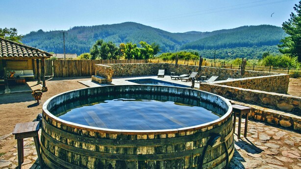 Piscina ao ar livre rústica com paredes de pedra e bancos de madeira, cercada por um pátio de pedra e vista para uma cadeia montanhosa coberta por floresta.