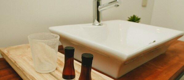 Un baño con un lavamanos blanco, un vaso de agua y dos botellas marrones sobre una superficie de madera.