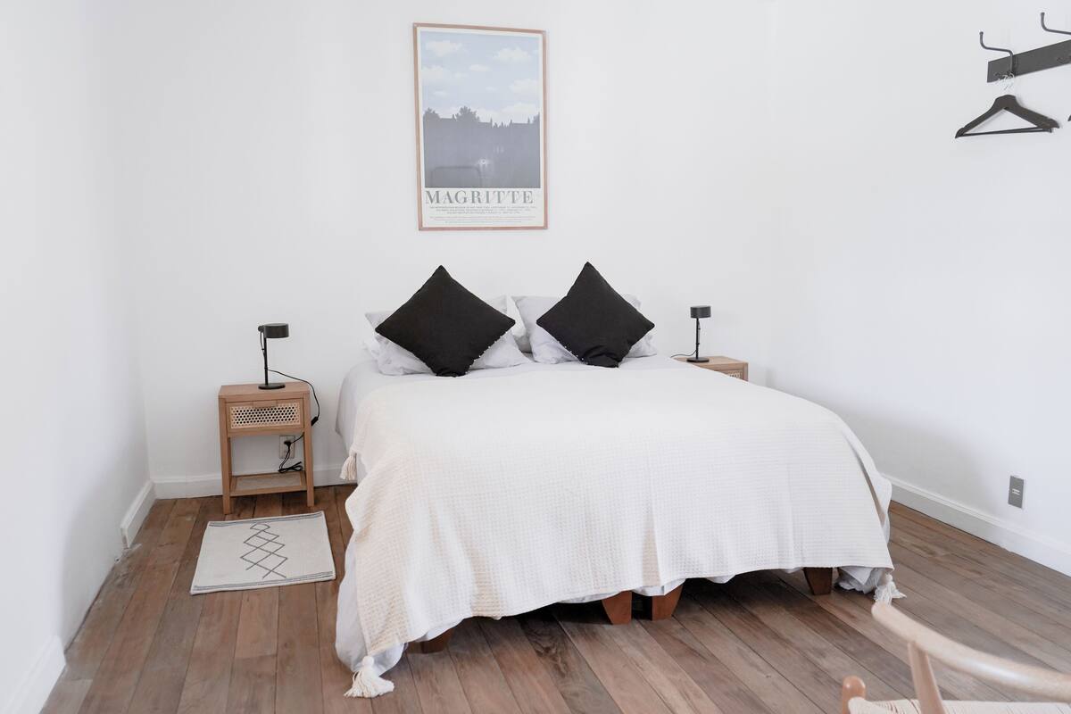 Ein Schlafzimmer mit einem großen Bett, zwei Nachttischen, einer Pendelleuchte, einem Poster an der Wand und Holzboden.
