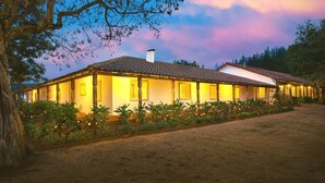 Um edifício de dois andares com telhado de telhas e uma varanda, rodeado por vegetação e uma árvore.