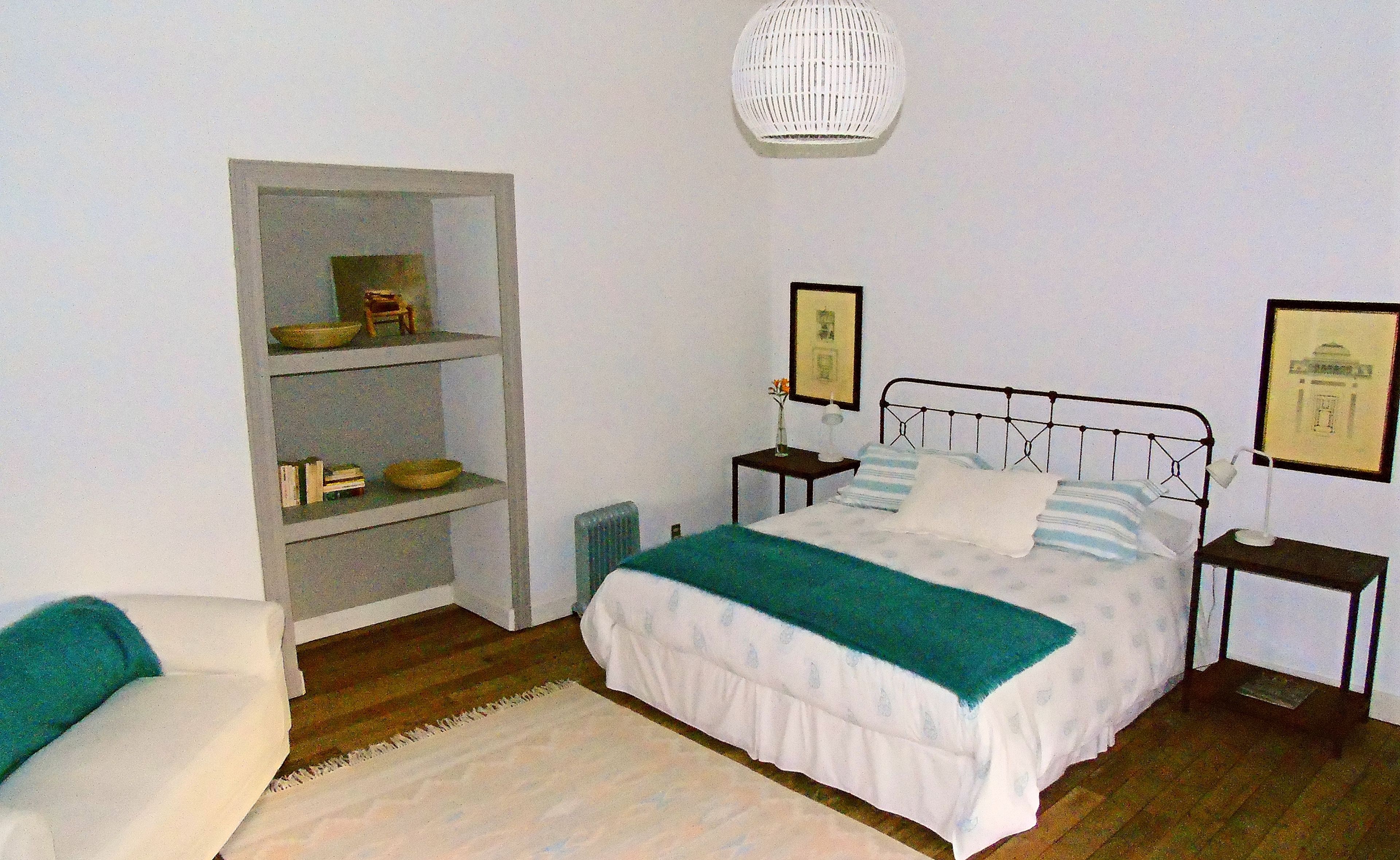 A bedroom with a bed, a nightstand, a bedside table, a shelf with books and a bowl, and a hanging light fixture.