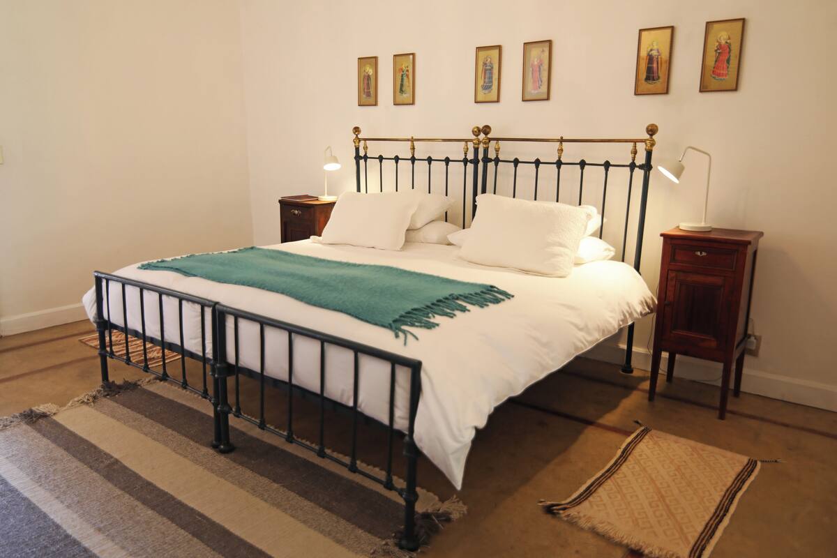 Ein ordentlich gemachtes Bett mit einer grünen Tagesdecke, ein Nachttisch mit einer Lampe und ein kleiner Holzbeistelltisch.
