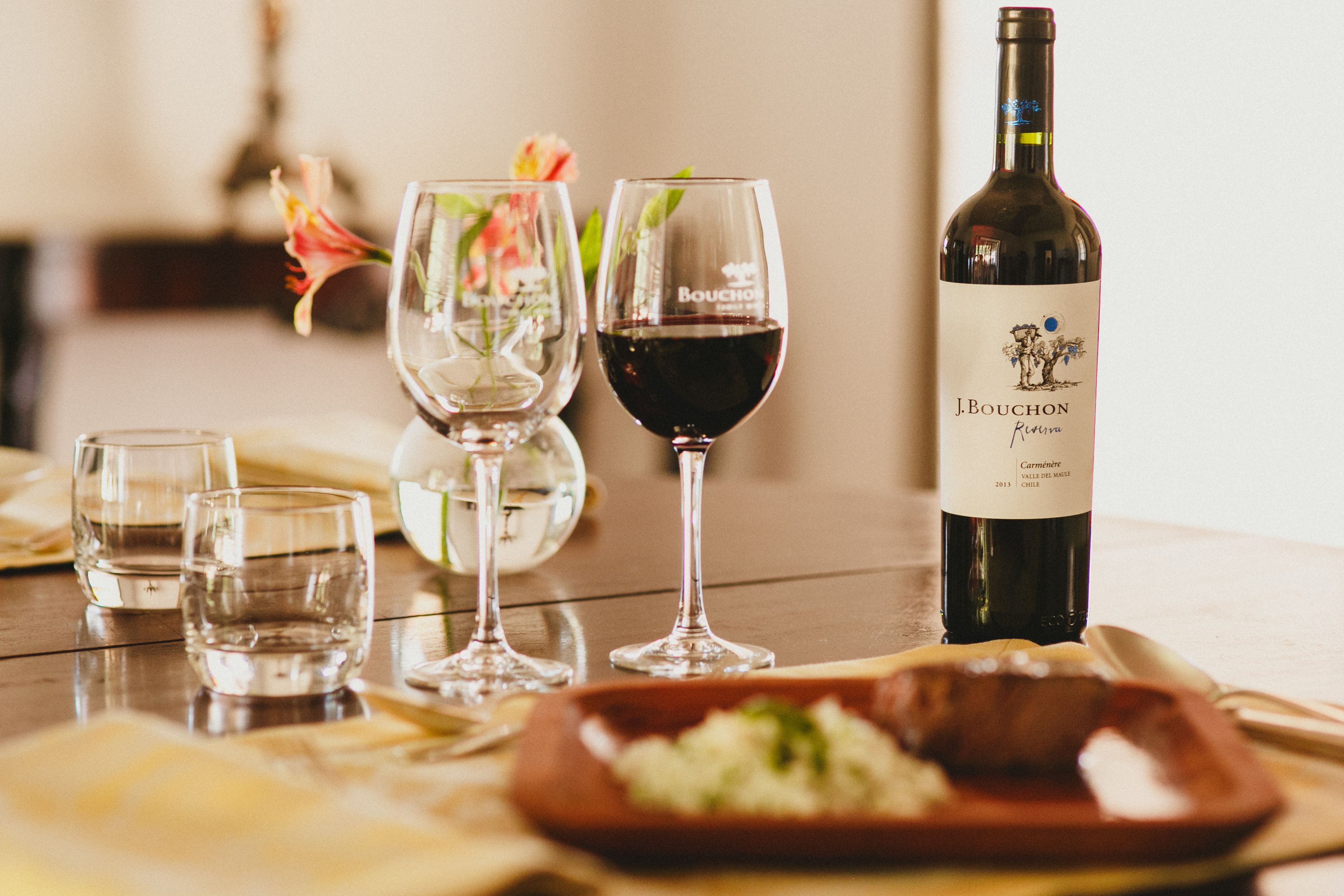 A dining table with a bottle of J. Bouchon wine, two wine glasses, and a plate of food.