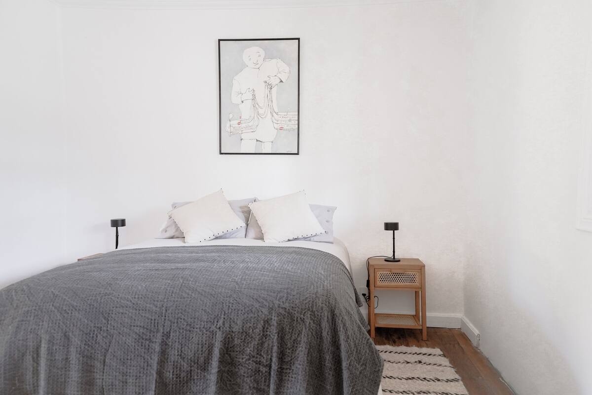 Ein Schlafzimmer mit Bett, Kissen, Nachttisch, Lampe und einem gerahmten Kunstwerk an der Wand.