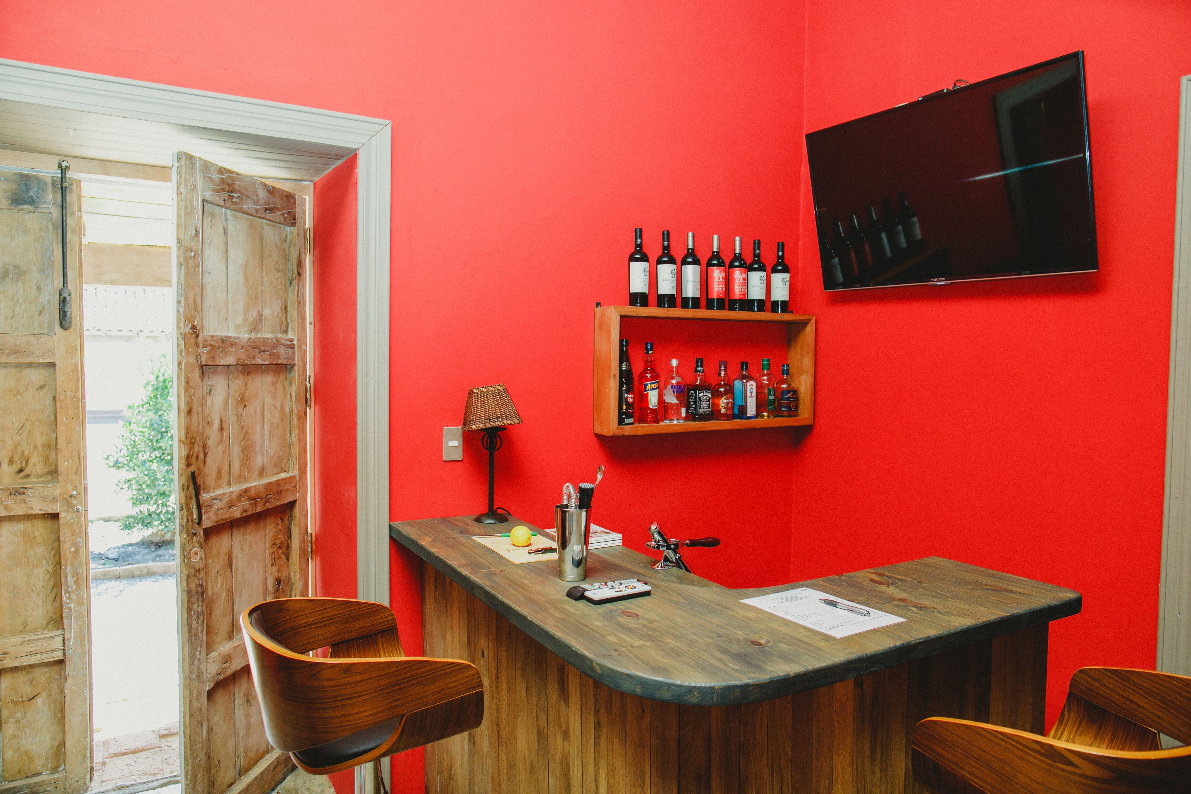 A bar area with a wooden counter, two wooden chairs, a TV mounted on the wall, and a shelf with bottles.
