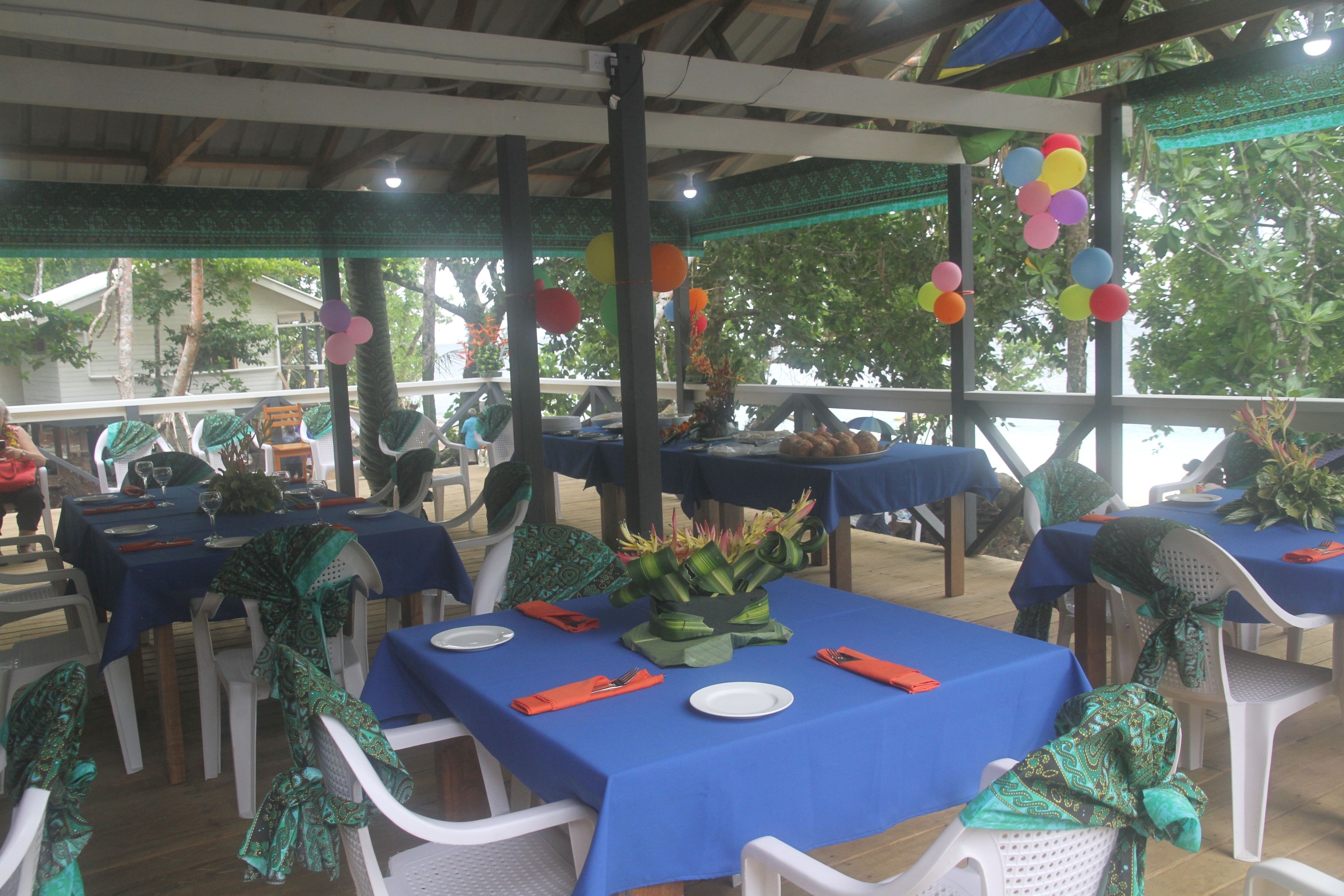 An outdoor dining area with tables covered in blue tablecloths and white plastic chairs, some with green fabric tied around the backrests.
