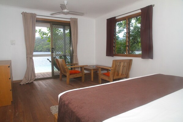 A room with a bed, a wooden table, two chairs, a window with curtains, and a view of a body of water. - Evis Resort (Halisi)