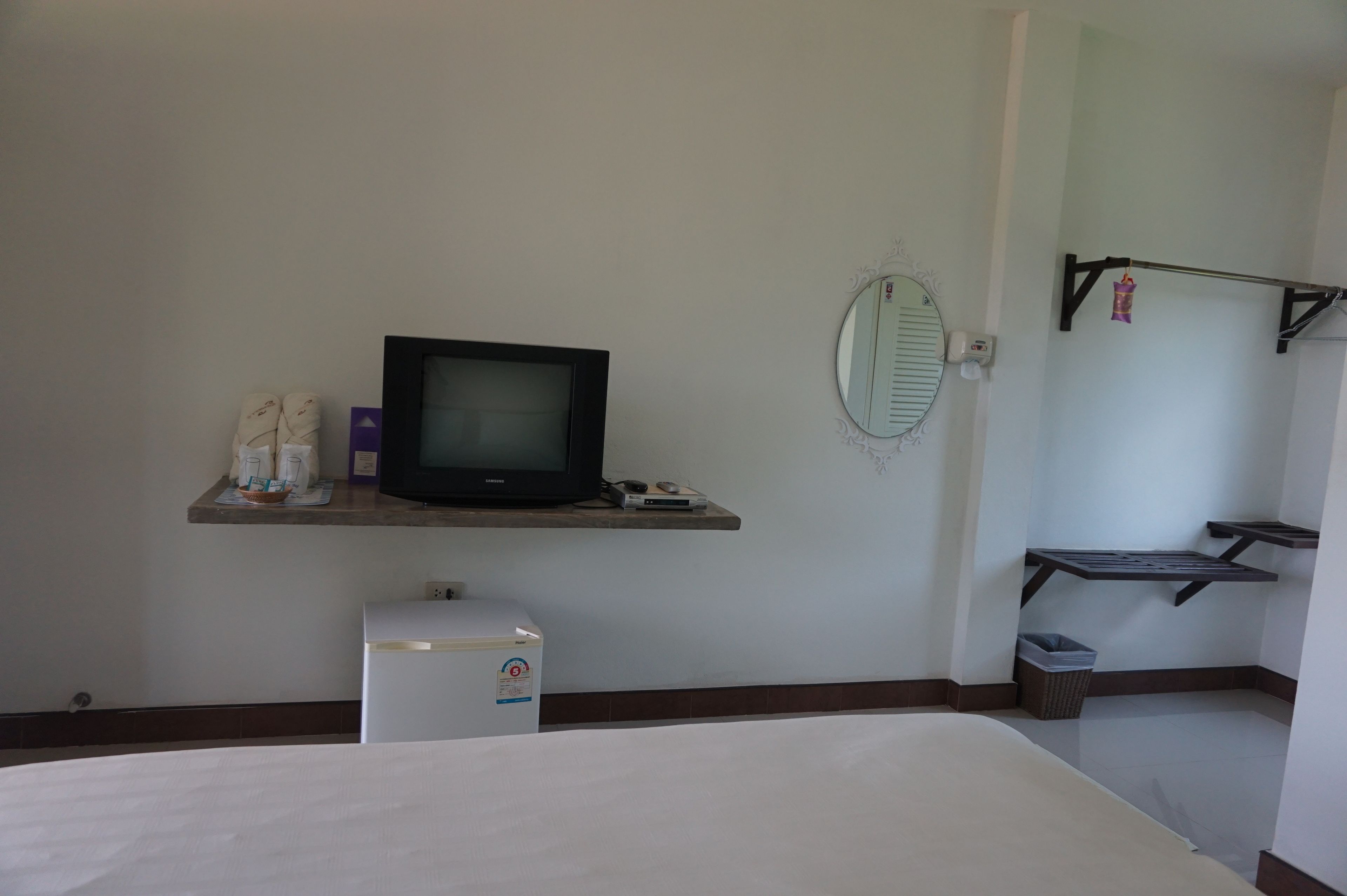 A hotel room with a bed, a television on a shelf, a small refrigerator, a trash bin, and a mirror on the wall.