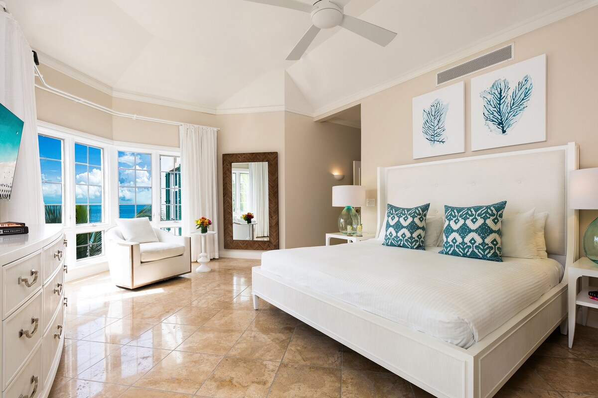A spacious bedroom with a large bed, a sitting area with a chair, and a view of the ocean through large windows.