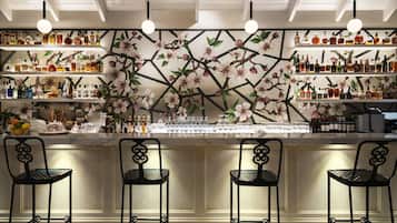 Zona de bar con un diseño floral en la pared, estantes con botellas y taburetes de metal negro.