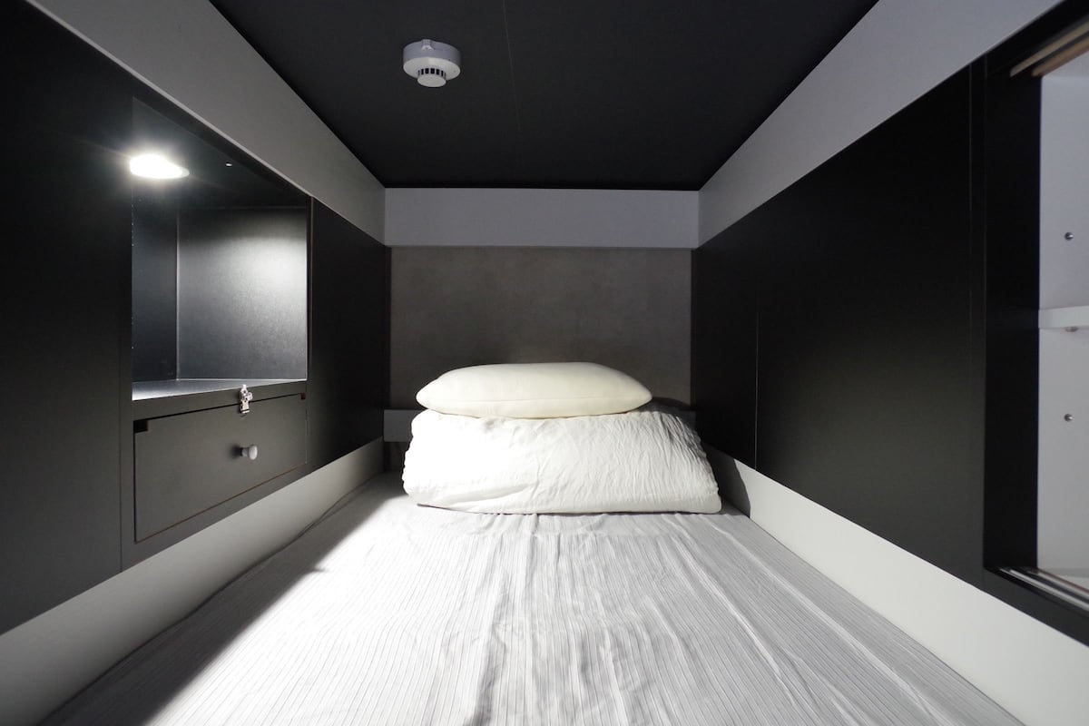 Una habitación estrecha, con poca luz, que tiene una cama individual, una mesita de noche y un espejo en la pared izquierda.