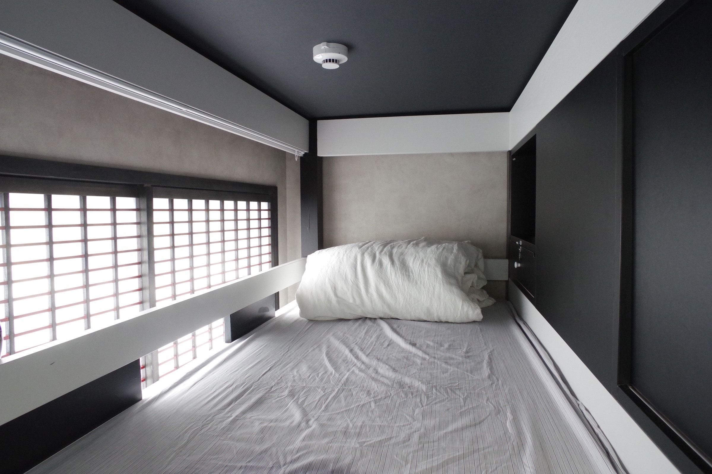 A small, modern room with a single bed, a window with blinds, and a ceiling light.